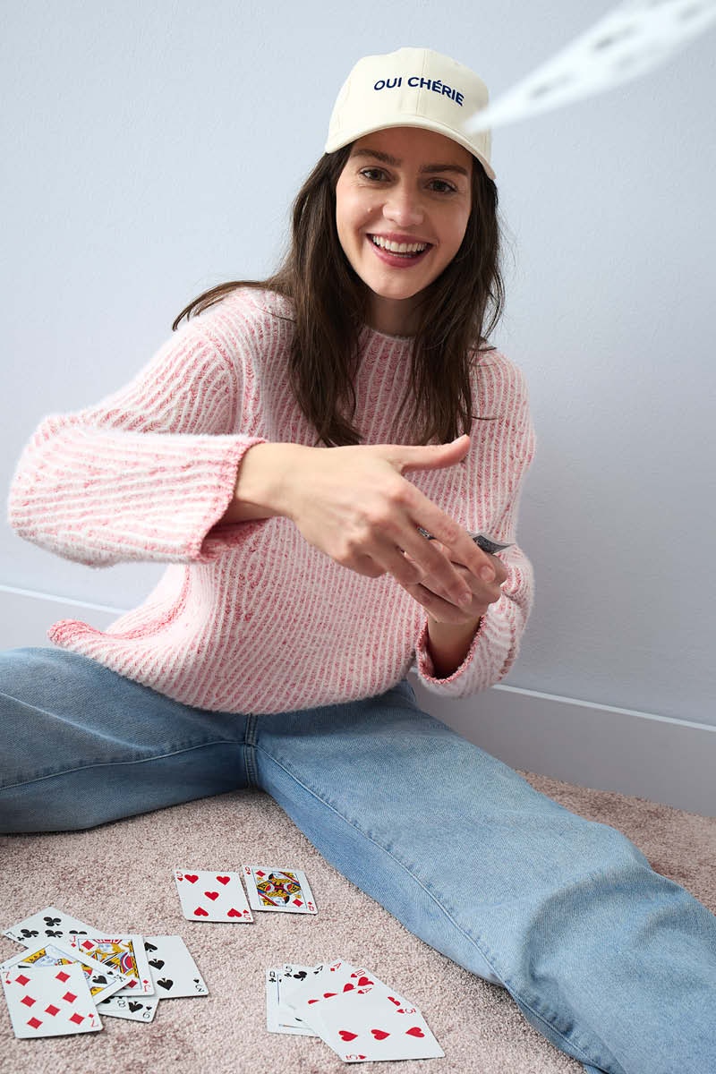 A woman sits on the floor and throws cards at the camera. She is wearing a pink jumper and light-coloured jeans.