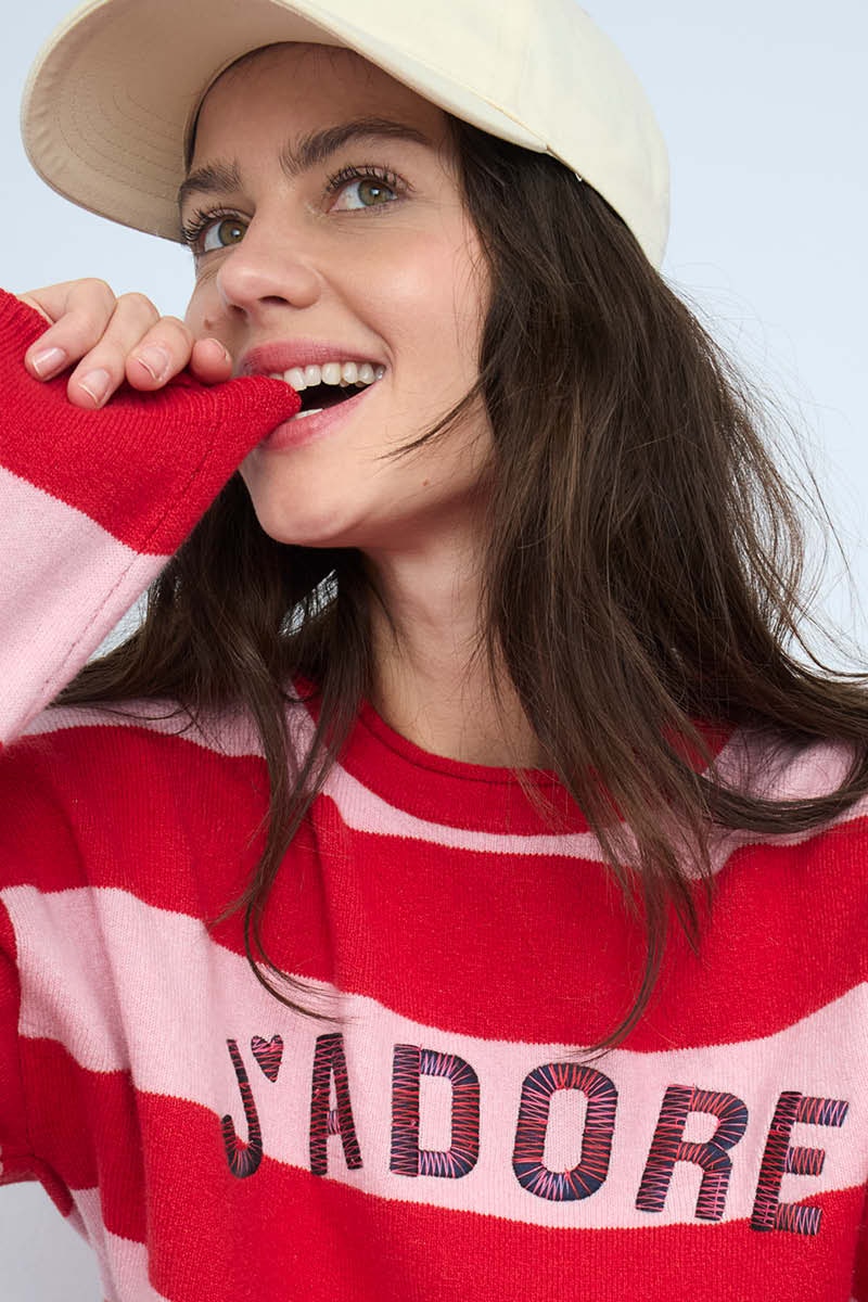 Close-up of a smiling woman wearing a red and pink striped jumper with the word 'Jadore' printed on the front.