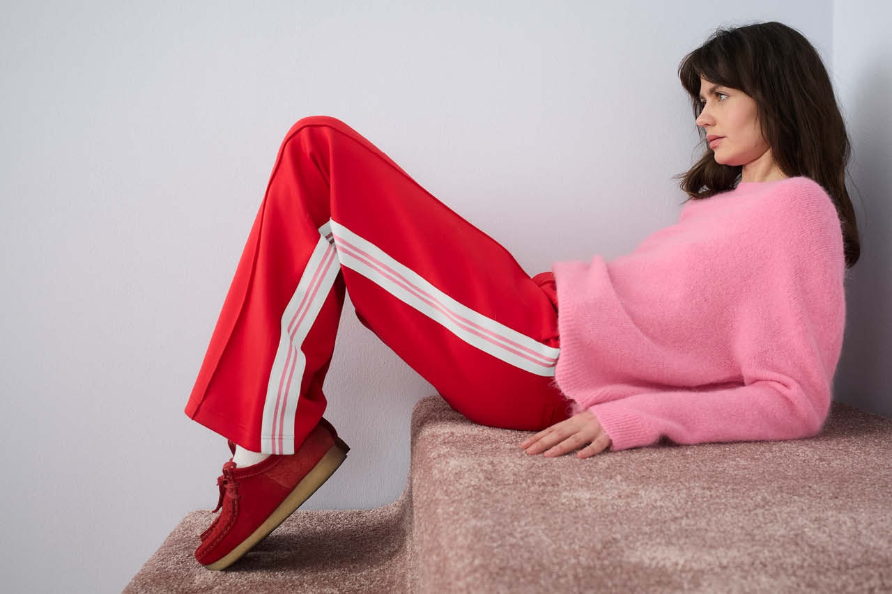 Woman sitting sideways on a plush carpet, wearing red trousers with contrasting white stripes down the sides and a pink jumper.