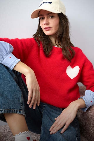 Woman wearing a red knitted jumper with a white heart motif on the chest, underneath which you can see a blue blouse with heart embroidery, and she is wearing jeans with it.