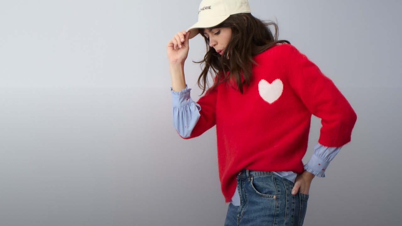 Frau mit einem roten Strickpullover und einem weißen Herzmotiv auf der Brust, darunter sieht man eine blaue Bluse mit Herzstickereien und sie trägt eine Jeans dazu.