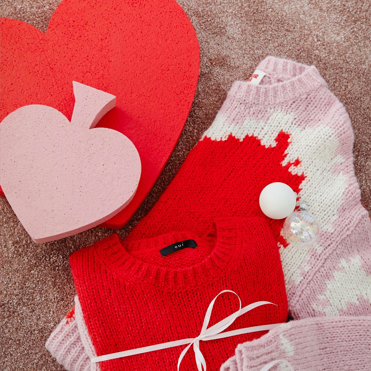 Zwei am Boden gefaltete Pullover einmal halbiert gelegt ein rosa PUllover mit roten und weißem Muster, darauf ein zusammengelegter roter Pullover. Links oben liegt ein roter Herzkarton und darauf ein Pic - Form Karton in rosa.