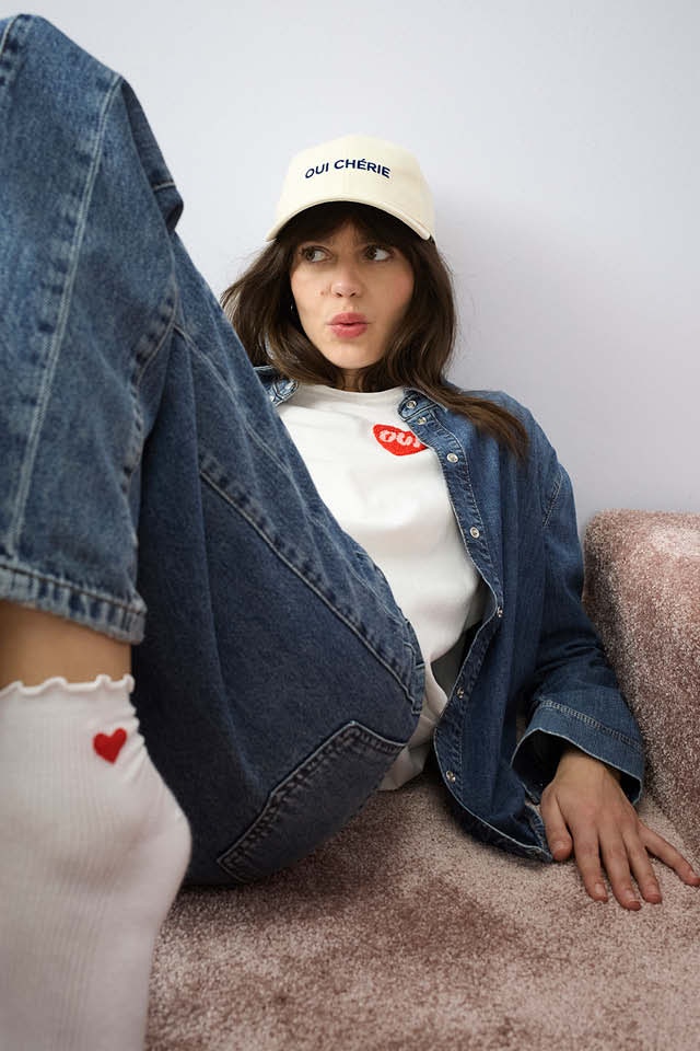 A woman is lying on the floor. She is wearing an open denim shirt with a white shirt with a red heart on the chest and matching denim trousers.