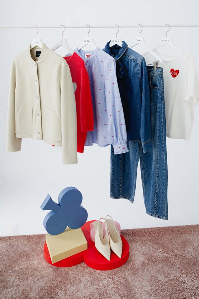 Eine Kleiderstange auf der ein roter Strickpullover mit einem weißen Herz auf der Brust hängt, dahinter eine gestreifte hellblaue Bluse, dann eine Jeansbluse, eine Jeanshose und ein weisses T-Shirt mit einem roten Herz auf der Brust.