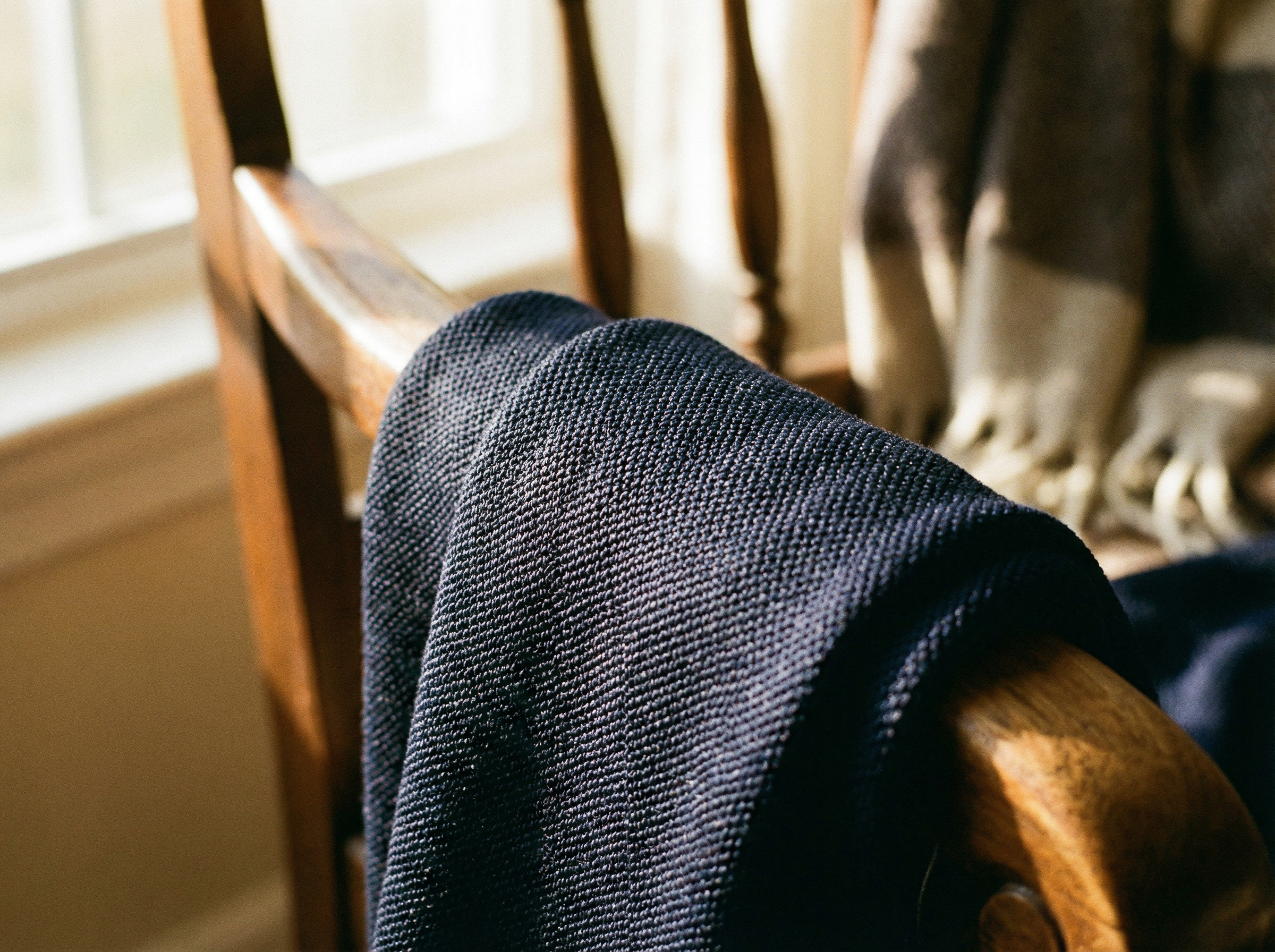 Ein blauer Strickpullover in Milano gestrickt hängt über einer Holzstuhl-Lehne mit Sonnenschein Reflektionen.