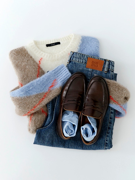 Gefalteter Pullover mit Karomuster in Weiß, Hellblau, Braun und Rot, kombiniert mit gelegter Jeans und braunen Loafern. 