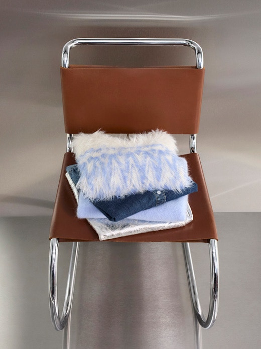 Auf einem Modernen Stuhl liegt ein Kleiderstapel, obenauf ein plüschiger Pullover in weiß mit hellblau gemustert, darunter sieht man eine Jeansbluse und weitere Kleidung in hellblau, grau und weiss.