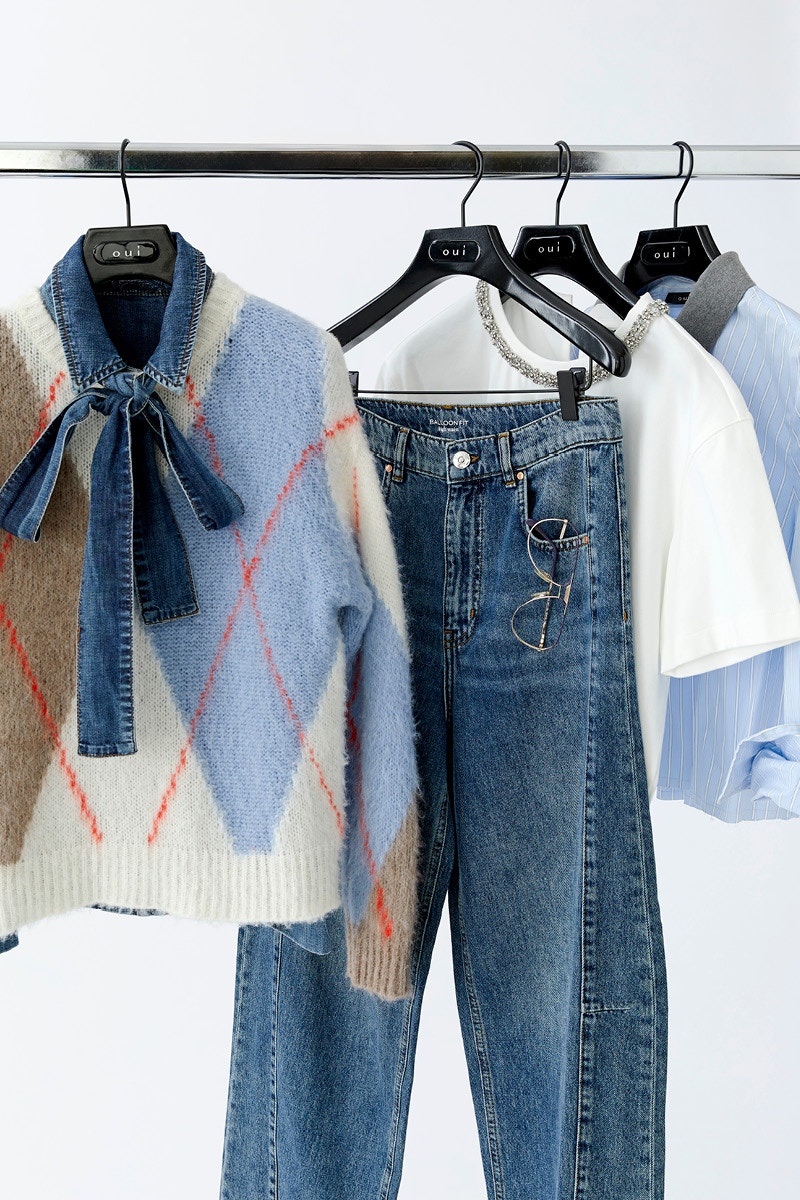 Mehrere Oui-Looks auf einem Kleiderständer: ein Argyle-Strickpullover mit Denim-Schleifenkragen, eine Balloon-Fit-Jeans mit Brille in der Tasche, ein weißes T-Shirt mit Glitzerkragen und eine hellblau gestreifte Bluse.