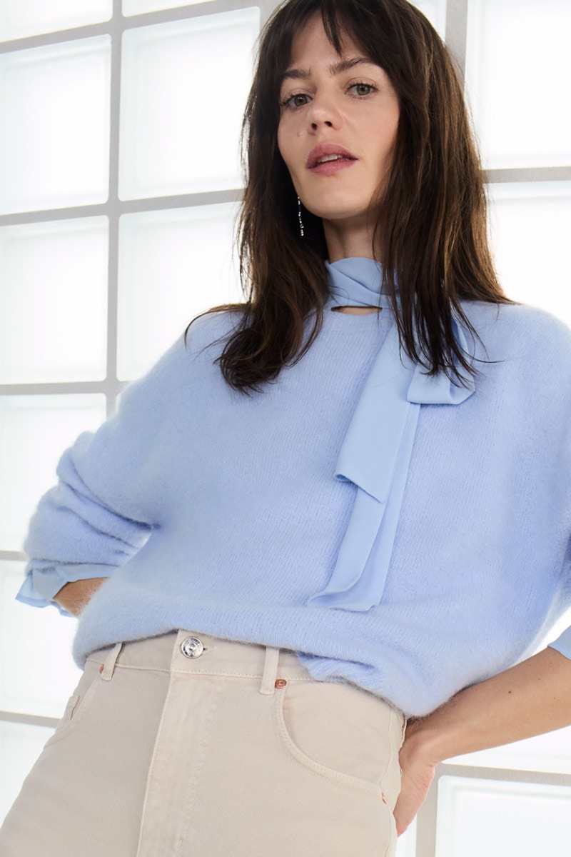 A woman wears a light blue, fluffy jumper with a tie bow at the neck, underneath a light blue blouse. She pairs it with beige high-waisted trousers.