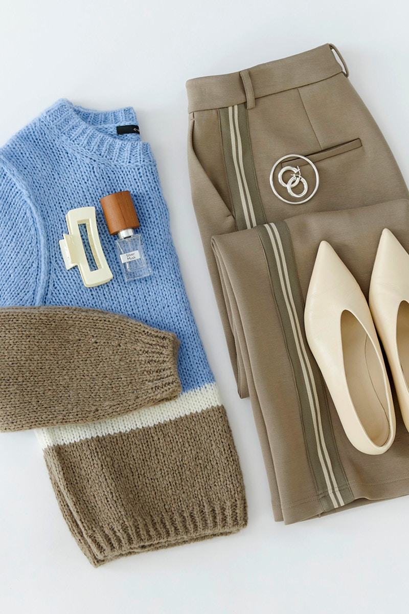 A folded knitted jumper in light blue, white and brown next to brown trousers with stripes down the sides. There are also cream-coloured court shoes, silver hoop earrings, a hair clip and a small bottle of perfume.