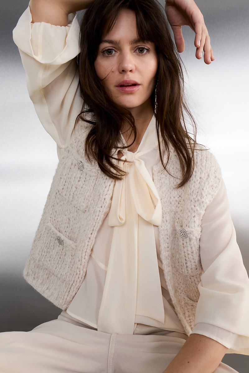 A woman wears a cream-white chiffon blouse with a long tie bow, topped with a textured, sleeveless knitted waistcoat with buttons. She pairs this with light-coloured, matching trousers.