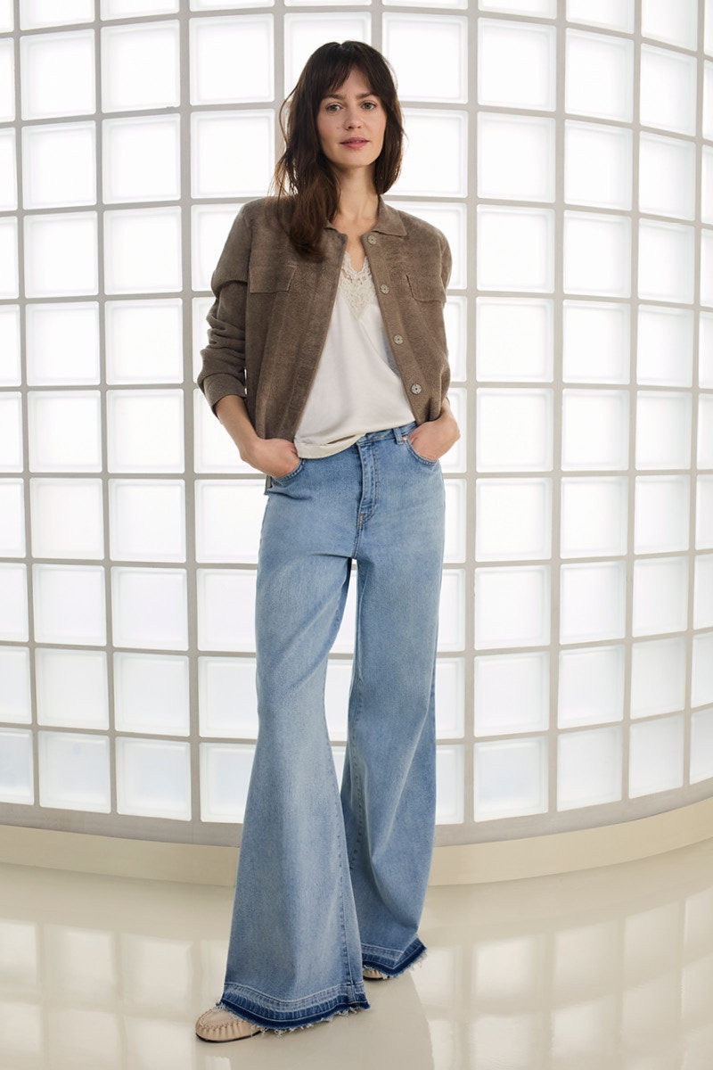 A woman wears a brown, casual shirt jacket over a white top with lace trim, pairing it with light blue, wide-leg jeans and flat beige shoes.