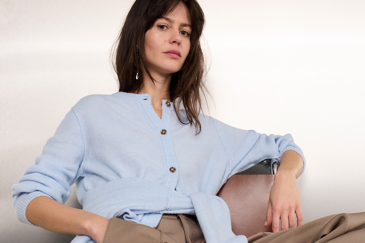 Woman sitting on a chair wearing a light blue cardigan with a light blue jumper tied around her waist, paired with brown trousers.