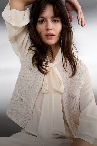 Eine Frau trägt eine cremeweiße Chiffonbluse mit langer Bindeschleife, darüber eine strukturierte, ärmellose Strickweste mit Knöpfen. Dazu kombiniert sie eine helle, passend abgestimmte Hose.