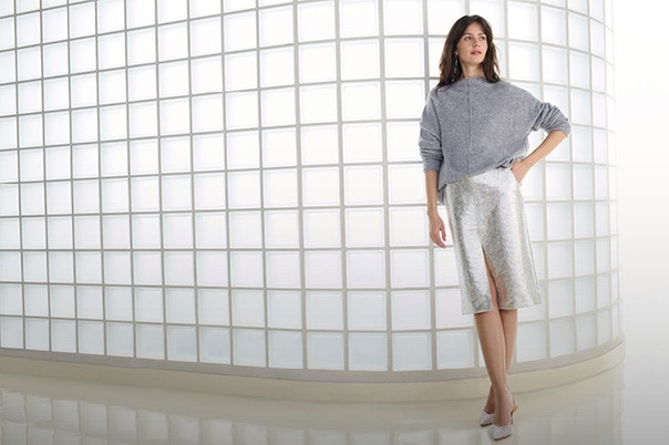 Eine Frau trägt einen grauen, lässigen Strickpullover, kombiniert mit einem silbernen, knielangen Rock mit Frontschlitz und glitzernden Pumps.