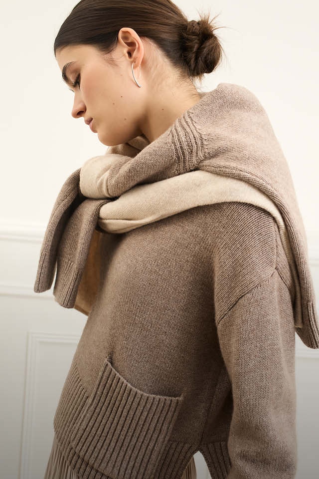 The woman is wearing a light brown knitted jumper and two more jumpers in beige and cream around her shoulders.