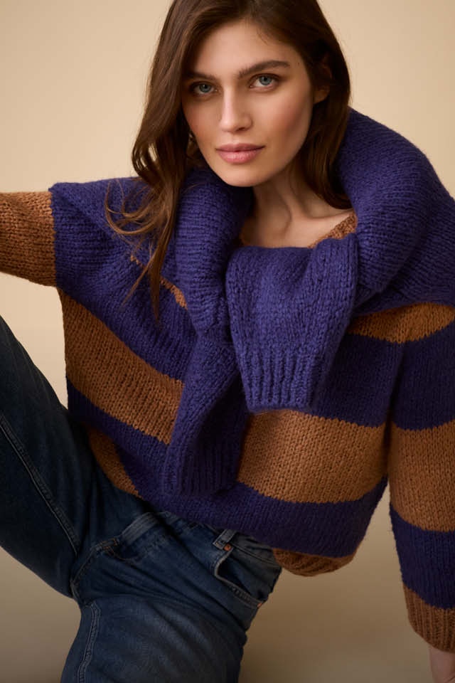 The woman is wearing a blue and brown striped jumper and has a blue jumper draped over her shoulders.