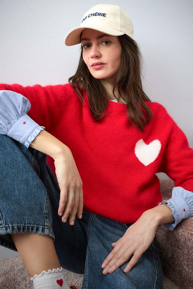 Woman wearing a red knitted jumper with a white heart motif on the chest, underneath which you can see a blue blouse with heart embroidery, and she is wearing jeans with it.