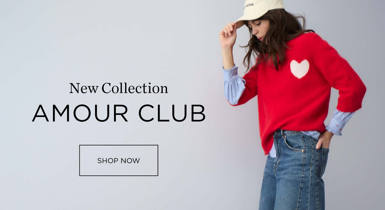 Woman wearing a red knitted jumper with a white heart motif on the chest, underneath which you can see a blue blouse with heart embroidery, paired with jeans. Text: New Amour Club collection with clickable area Shop now.