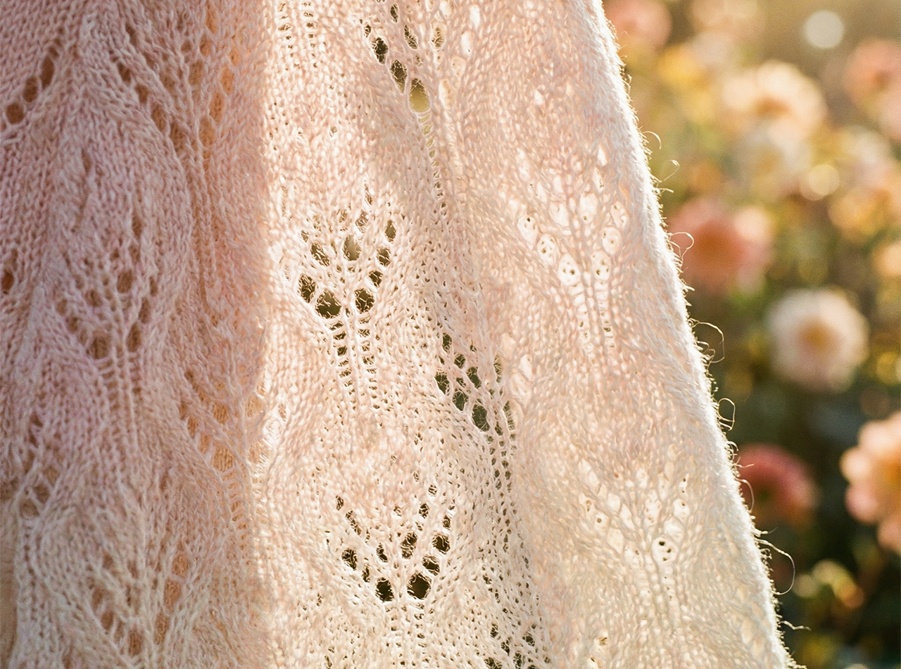 Detailaufnahme eines hellen, fein gestrickten Pullovers mit Pointelle-Muster, der im Sonnenlicht vor unscharfen Blumen im Hintergrund hängt.