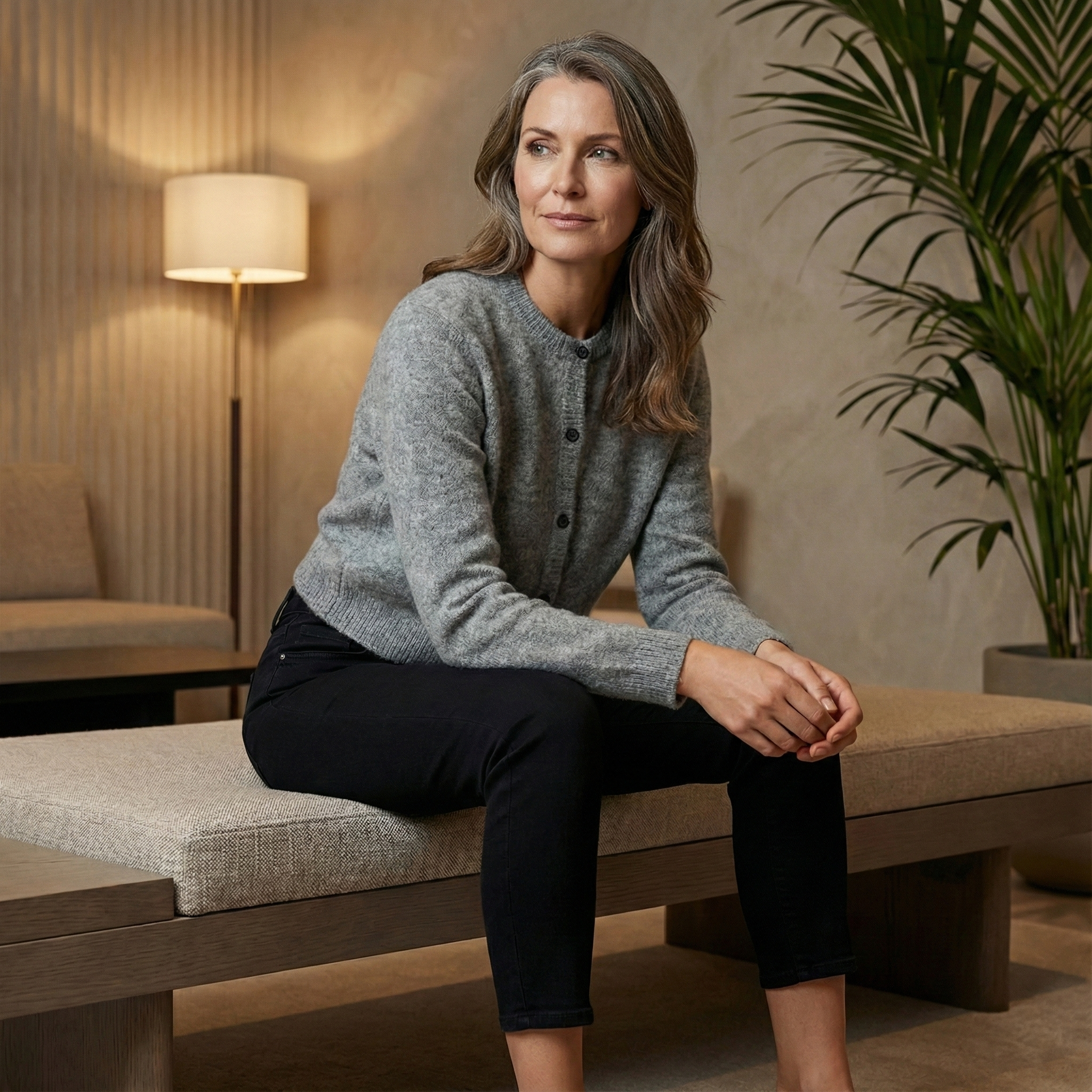 Eine Frau sitzt auf einer gepolsterten Bank, trägt eine graue Strickjacke mit Knöpfen und eine schwarze Hose.