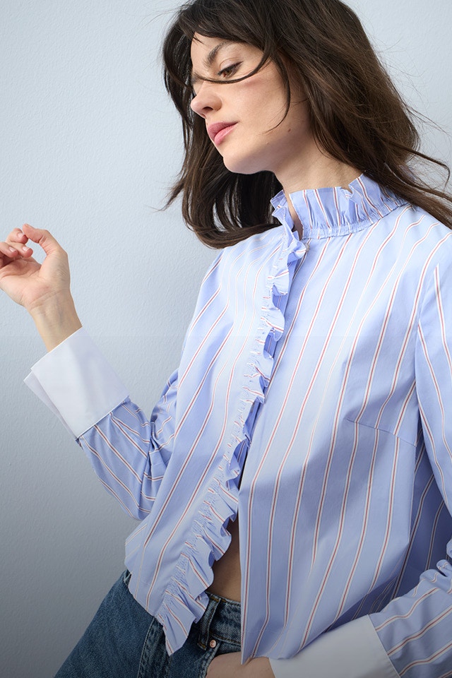 A woman wears a light blue striped blouse with ruffles on the collar and along the button placket, paired with jeans.