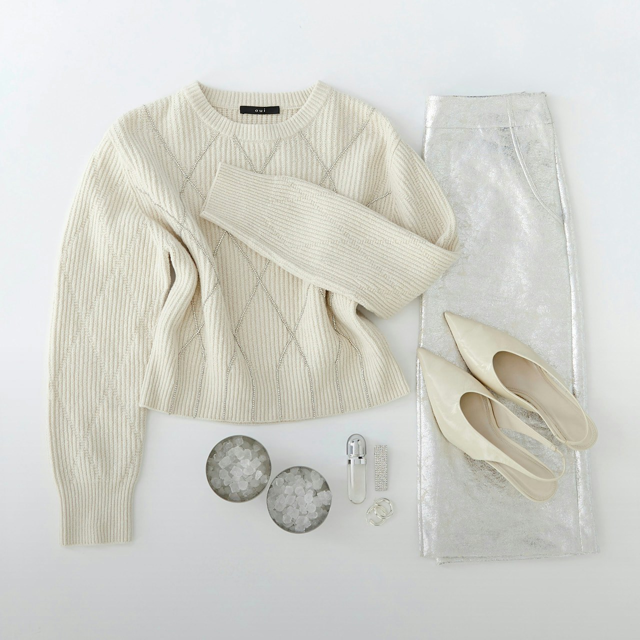 Ein cremefarbener Strickpullover liegt neben einem silbernen Rock, cremefarbenen Pumps und kleinen silbernen Accessoires.