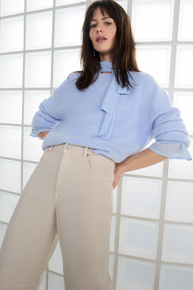 Eine Frau trägt einen hellblauen, flauschigen Pullover mit einer hellblauen Bluse darunter und der Bindeschleife am Hals. Sie kombiniert dazu eine beige High-Waist-Hose.