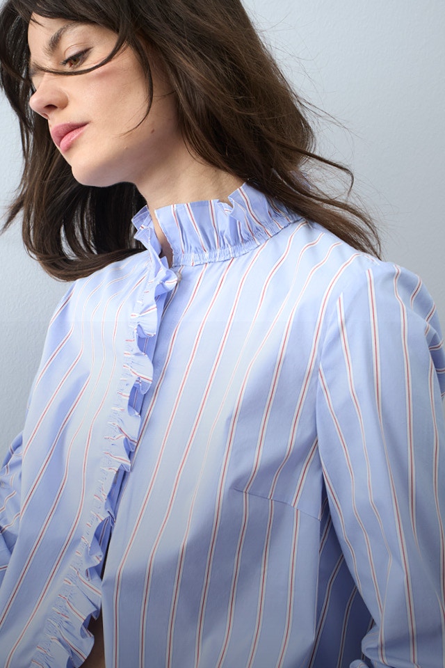 Eine Frau trägt eine hellblau gestreifte Bluse mit Rüschen am Kragen und entlang der Knopfleiste, kombiniert mit einer Jeans.