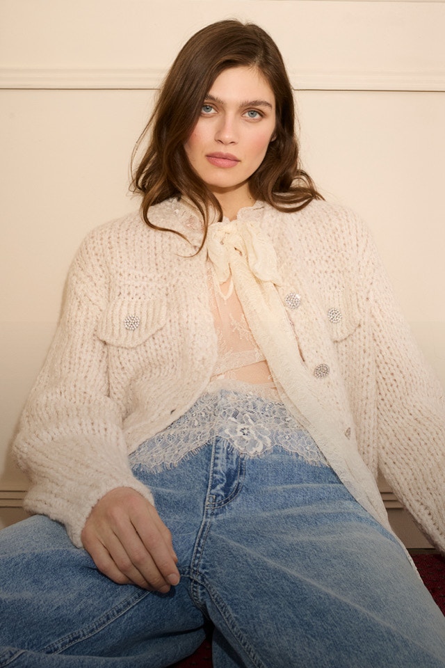 Frau sitzend trägt eine weiße Strickjacke mit Schmuckknöpfen und darunter eine Spitzenbluse leicht transparent mit einer Jeanshose.