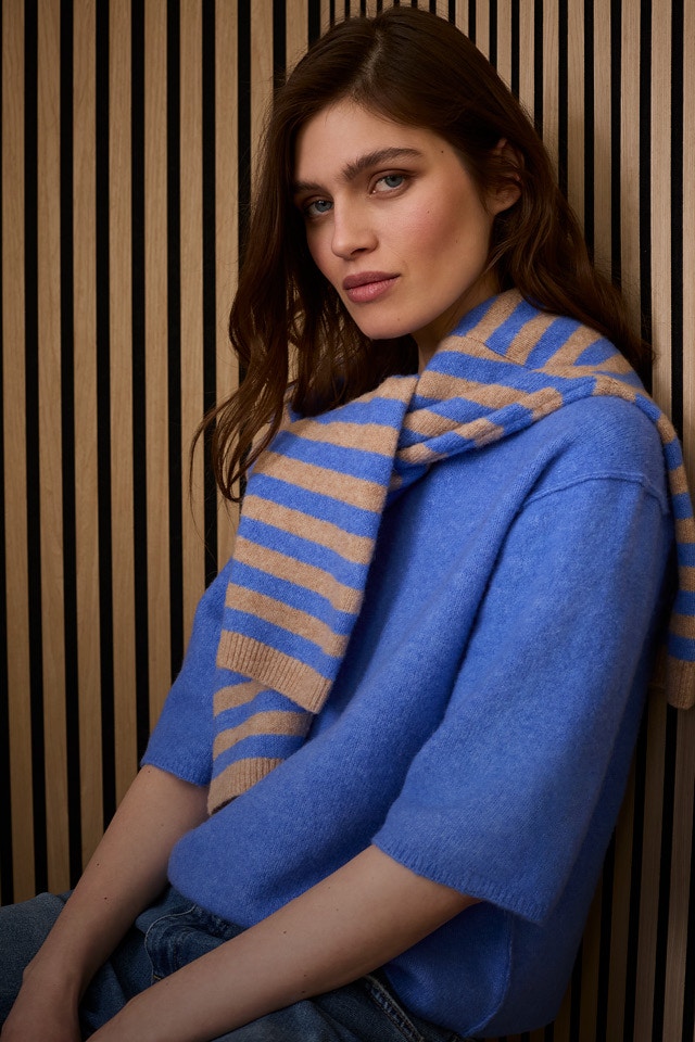 Frau mit blauem Pullover, über den Schultern eine Strickjacke gestreift aus blau und brauntönen gelegt mit eine Jeans.