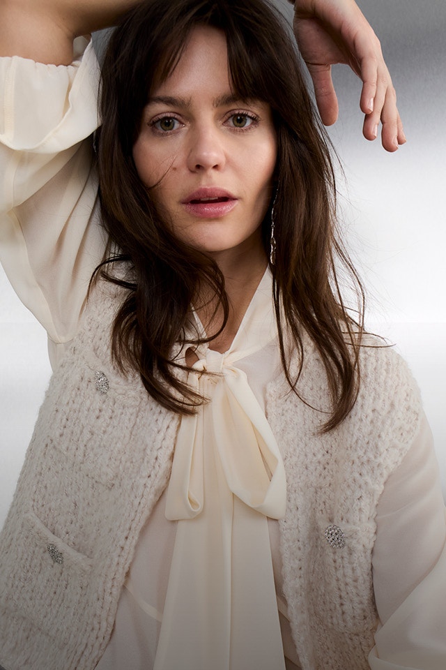 A woman wears a cream-white chiffon blouse with a long tie bow, topped with a textured, sleeveless knitted waistcoat with buttons.