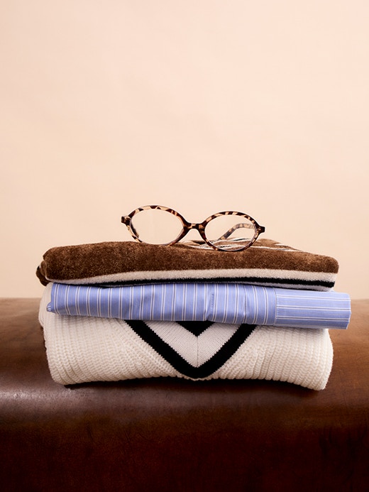 Ein Stapel Kleidung, unten ein weisser Pullover mit braunem V-Ausschnitt, darüber eine blau-weiss gestreifte Bluse und darauf ein Shirt in braun-weiss gestreift. 