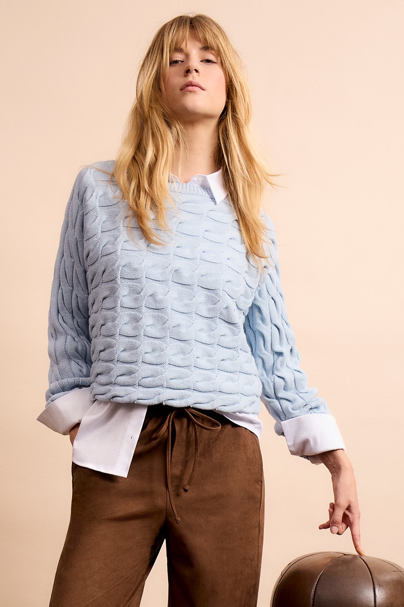 Frau mit hellblauem Pullover, darunter weisser Bluse und brauner Hose.