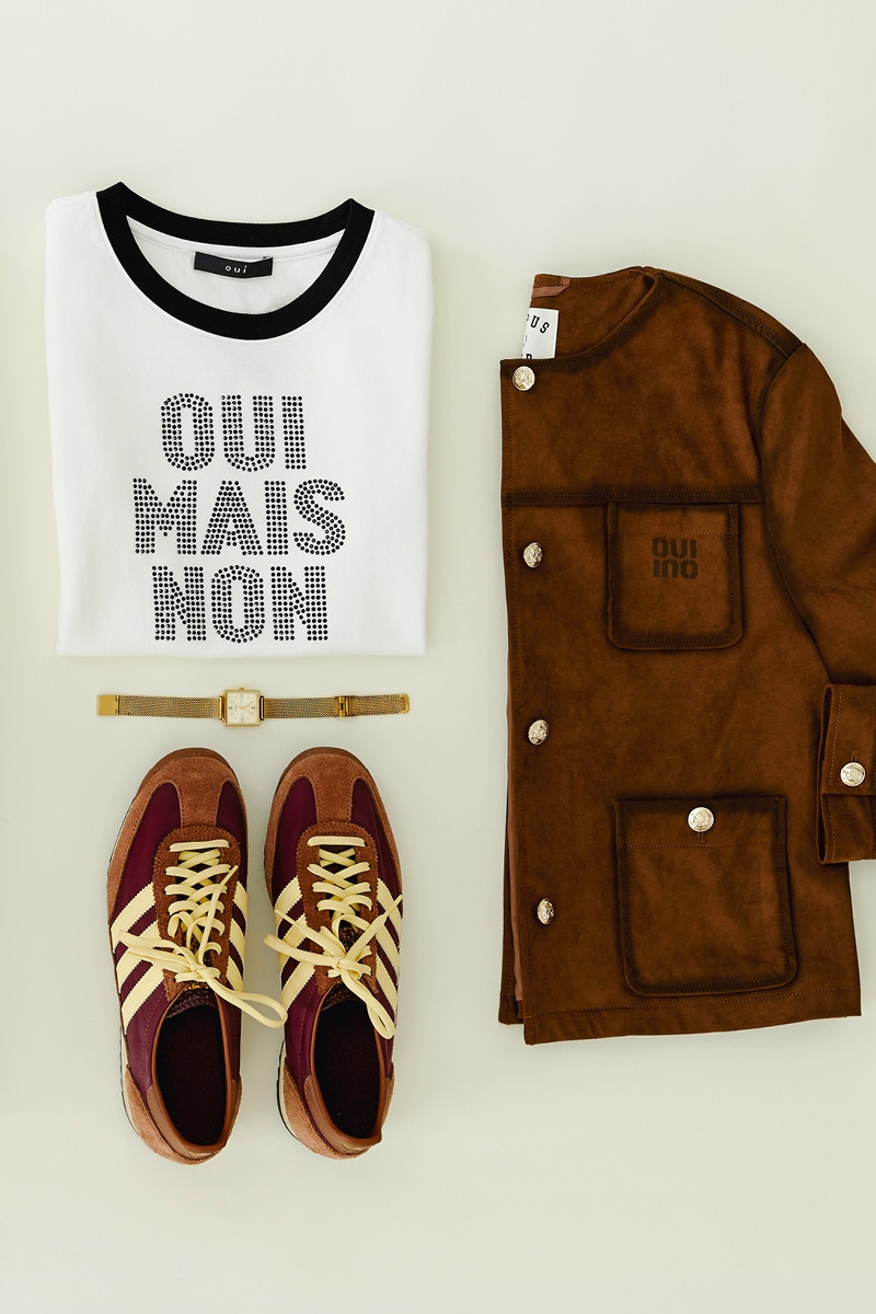 Gelegte Kleidung eine halbgefaltete braune Jacke recht, links oben ein gelegtes T-Shirt in weiß mit Schrift: Oui Mais Non in schwarzen Steinen, darunter braune Sneaker.