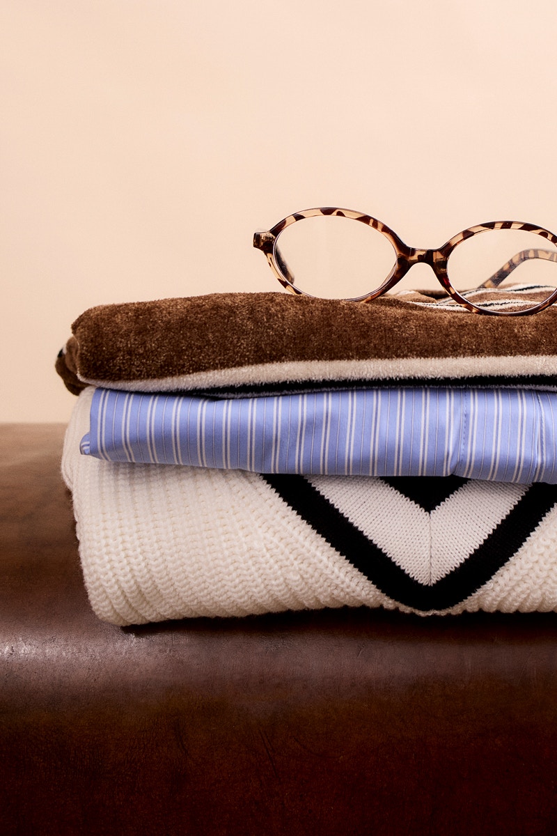 A pile of clothes, at the bottom a white jumper with a brown V-neck, on top of that a blue and white striped blouse and on top of that a brown and white striped shirt.