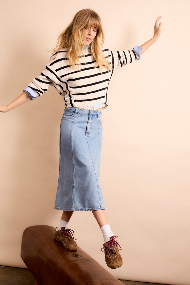 Woman wearing a beige and brown striped jumper, a blue and white blouse underneath, and a denim skirt.