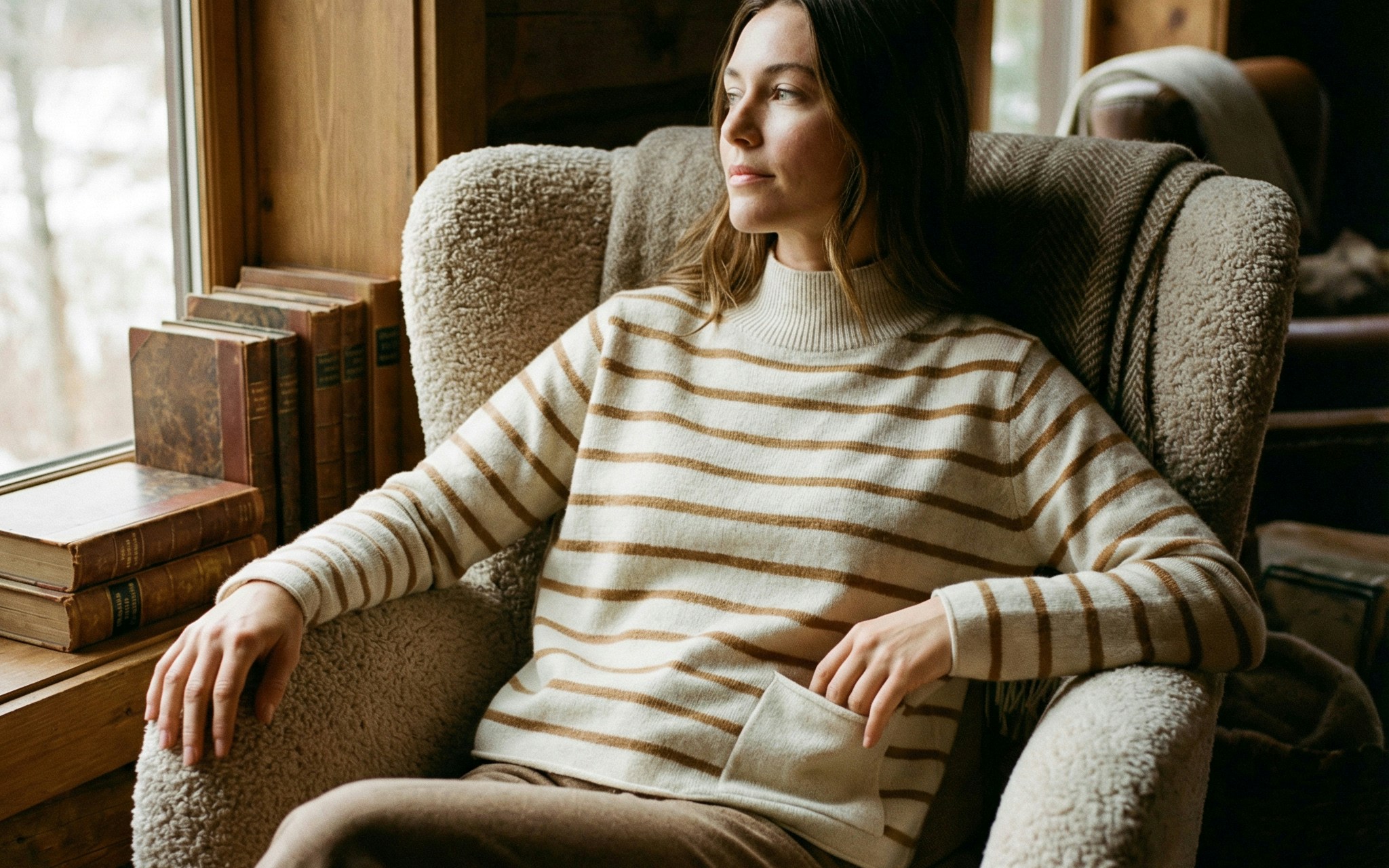 Woman sitting relaxed in a beige armchair, wearing a striped knitted jumper in beige and brown with a small pocket on the bottom right.