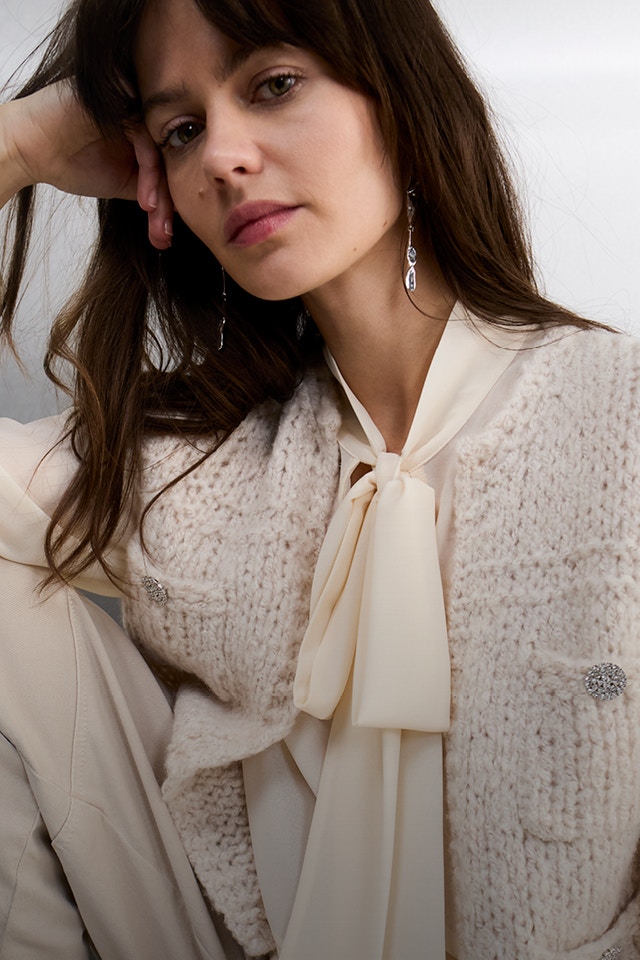 A woman wears a cream-white chiffon blouse with a long tie bow, topped with a textured, sleeveless knitted waistcoat with buttons.