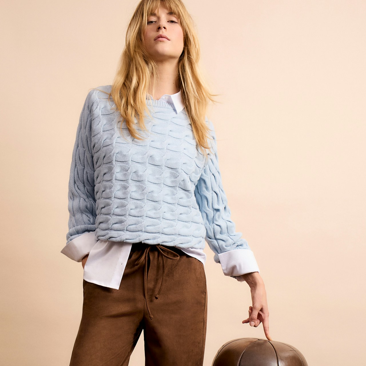 Woman wearing a light blue cable-knit jumper, white blouse underneath and brown trousers.