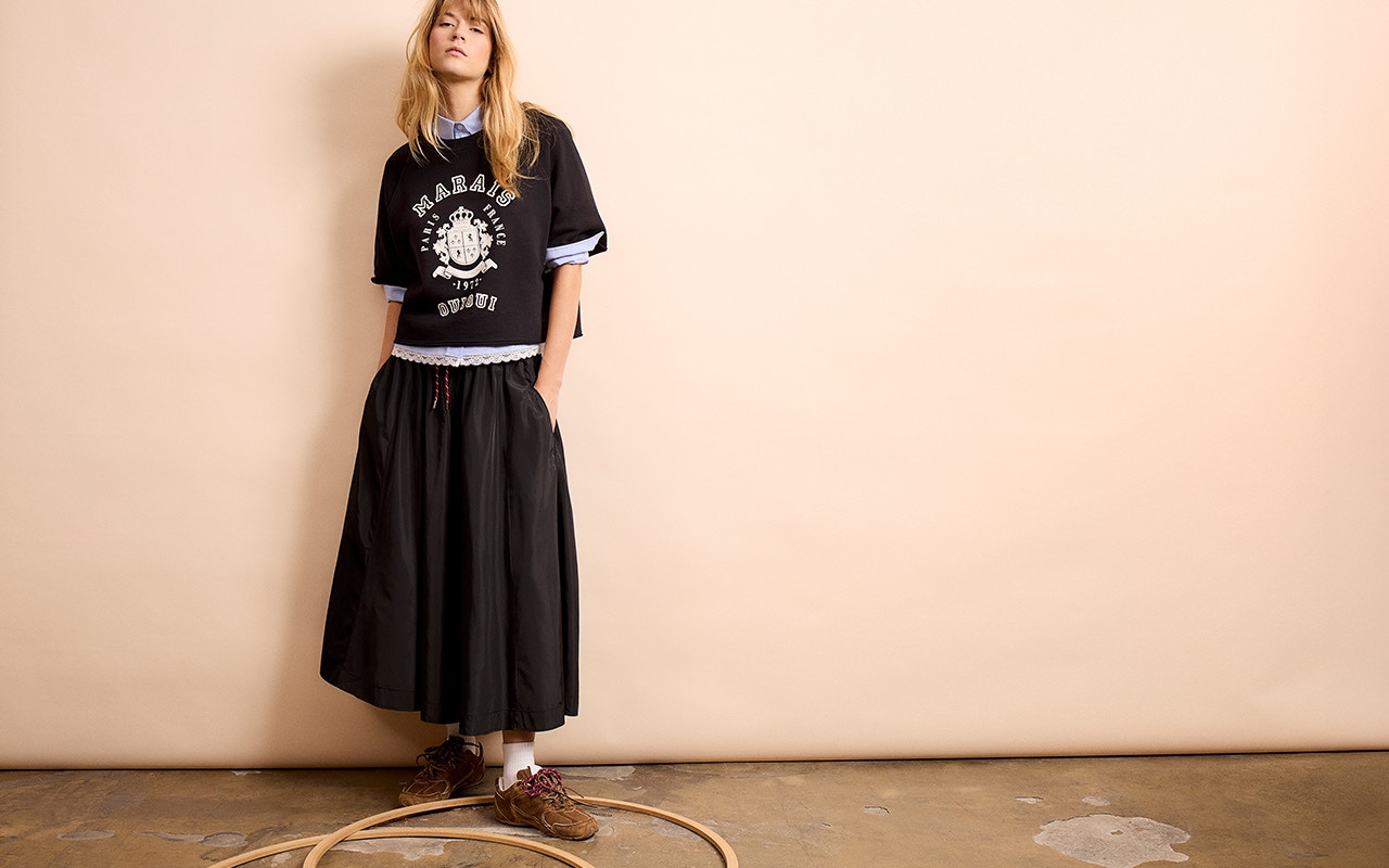 Frau mit schwarzem T-Shirt mit weißem Aufdruck, darunter eine blau-weiß gestreifte Bluse und ein schwarzer Rock.