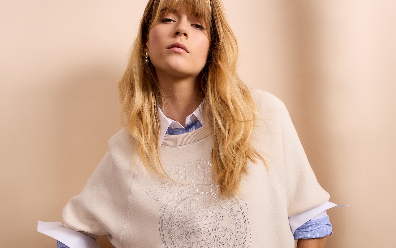 Woman wearing a beige jumper with a light-coloured print, underneath a blue and white striped blouse and jeans.