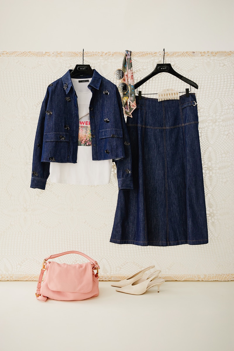 Stillleben mit Damenmode von Oui: Dunkelblaue Jeansjacke mit floralen Applikationen kombiniert mit weißem T-Shirt mit Print und dunkelblauem Jeansrock, präsentiert auf Kleiderbügeln. Feminines Denim-Outfit aus den Oui Damenkollektionen auf oui.com.