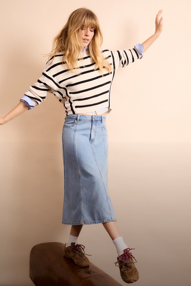 Frau mit gestreiftem Pullover in beige-braun, blau-weisser Bluse darunter und Jeansrock.