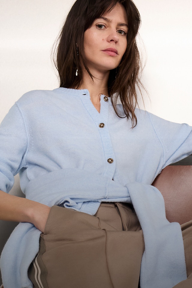 Frau trägt eine hellblaue BLuse um die Hüfte einen hellblauen Pullover gebunden und dazu eine braune Hose.