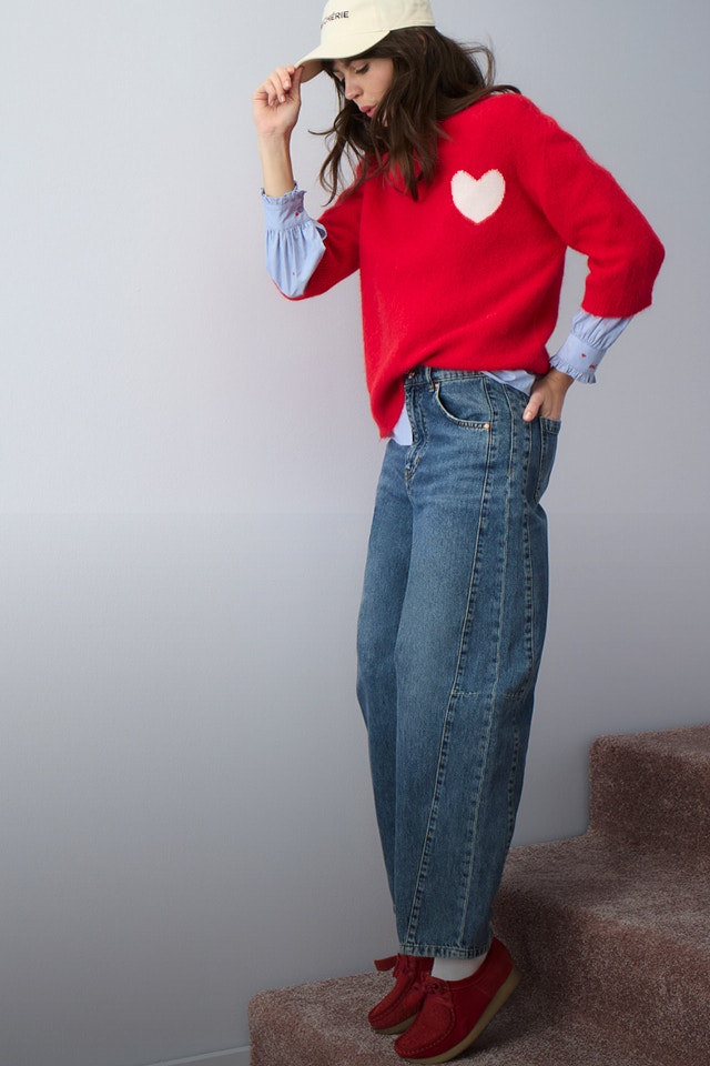 Woman wearing a red jumper with a white heart on the chest, underneath a striped blue blouse and jeans.