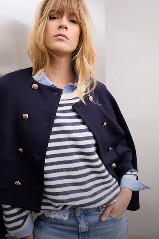 Frau trägt einen blau weiss gestreiften Pullover, eine blaue Jacke darüber und eine Jeans.