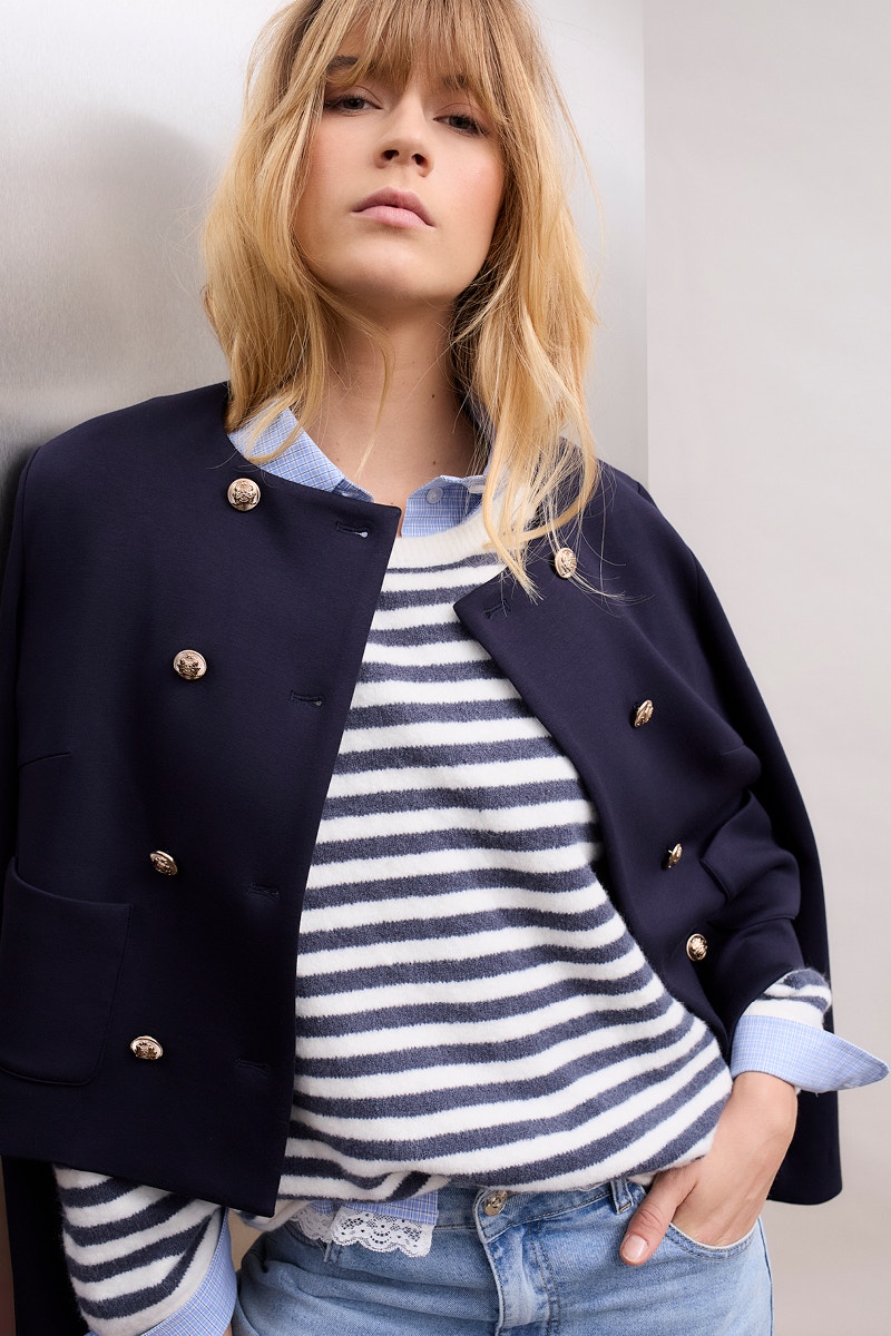 Frau trägt eine dunkelblaue Jacke, darunter ein blau-weiss gestreiften Strickpullover und eine Jeans.
