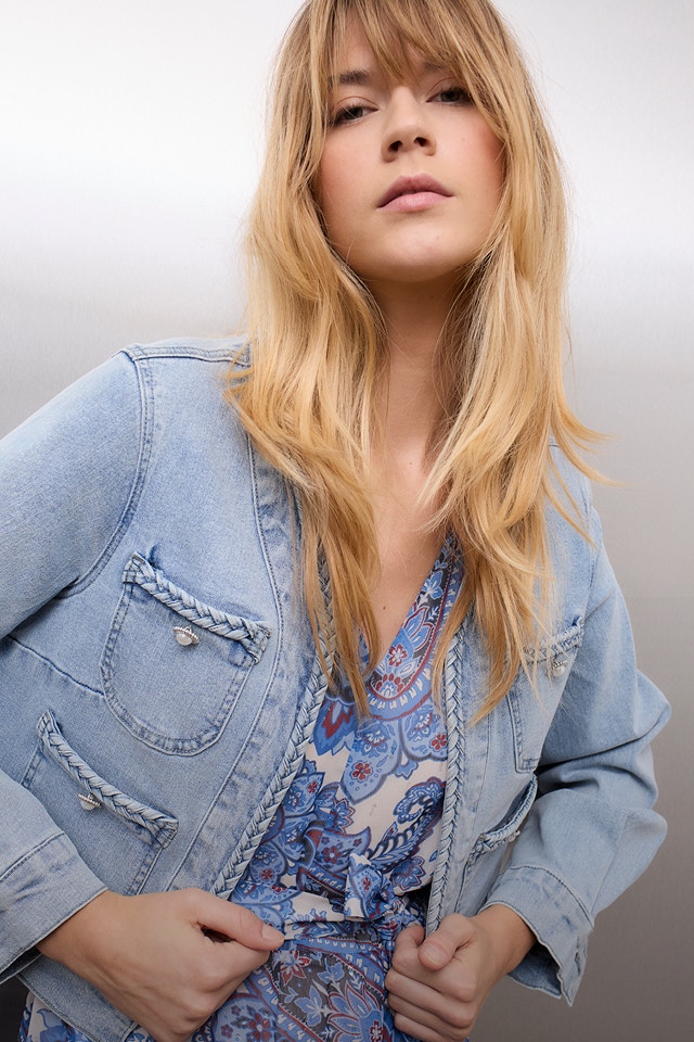 Woman wearing a denim jacket, underneath a white and blue paisley blouse and denim trousers.