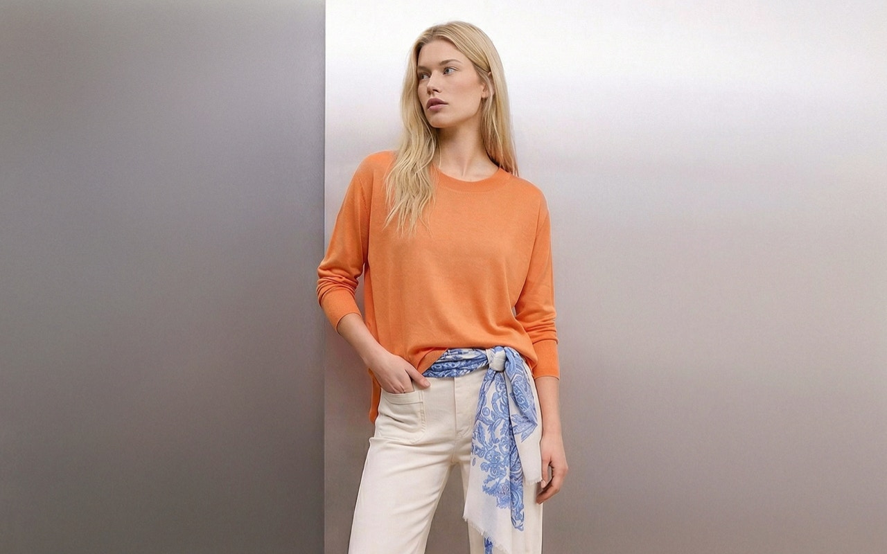 Frau mit orangem Pullover, blau-weissem Tuch als Gürtel in der weissen HOse.